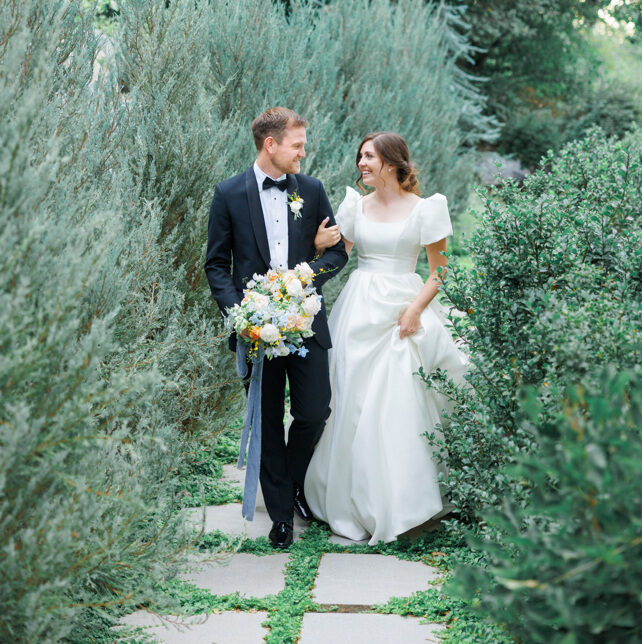 Happy couple walks outside surrounded by lush greenery, with the bride in a stunning modest wedding dress with puffed sleeves and a full skirt, and a bouquet with white, orange, and blue accents. Wedding inspiration #2024weddingtrends #Utaheventplanner #luxuryeventplanner #weddingtrends #weddingideas