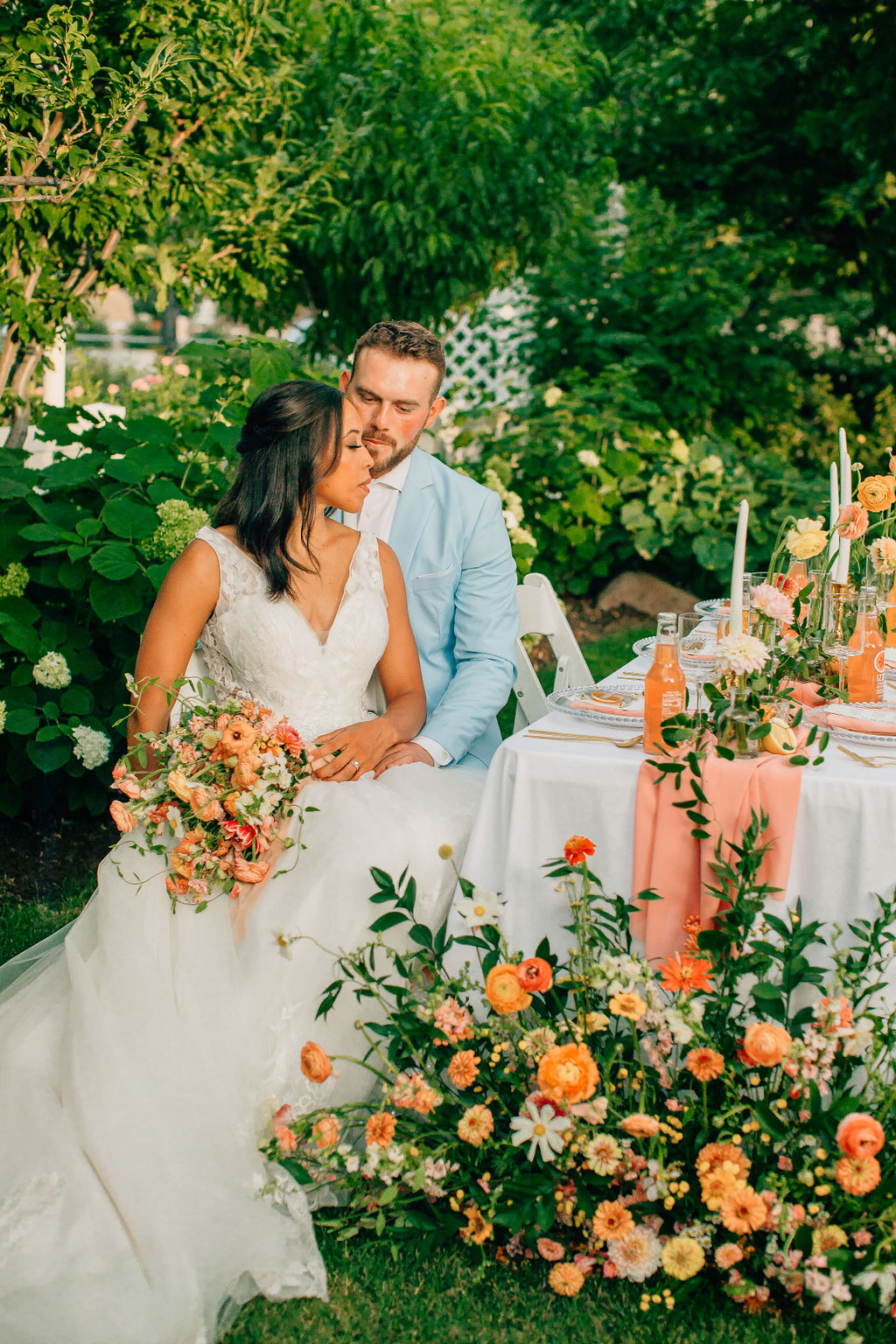 Bride and groom sit close alongside a citrus summer-inspired wedding tablescale filled with fruity peach pink and orange accents outside in a garden #citrusgardenwedding #gardenwedding #citrusweddingdesign #weddingdesignerslc