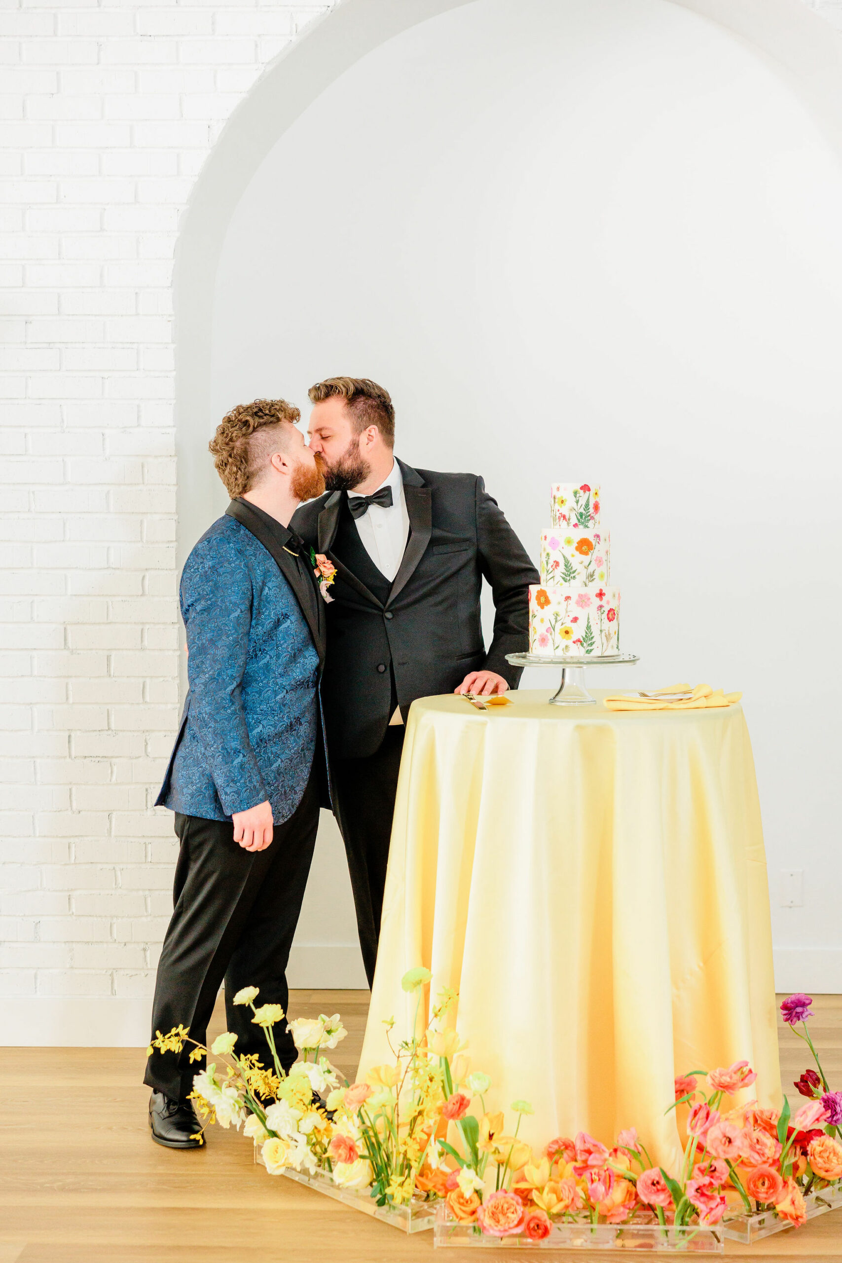 Couple kisses next to unique wedding cake with pressed flowers in shades of pink, orange, purple, yellow, and green. Nontraditional wedding cakes unique cake inspiration #2024weddingtrends #Utaheventplanner #luxuryeventplanner #weddingtrends #weddingideas